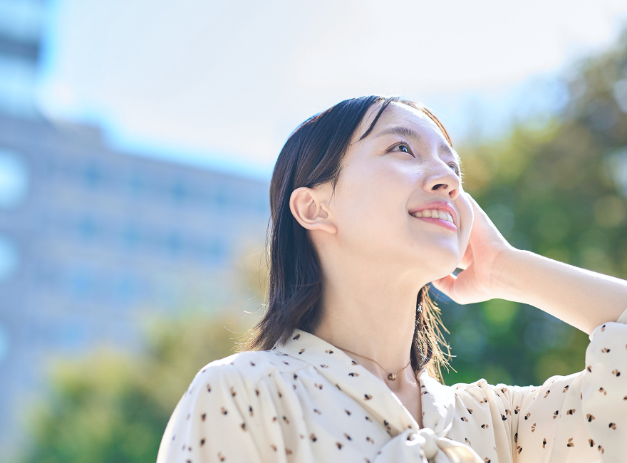 白地に小さな模様が入ったブラウスを着た女性が屋外でリラックスした表情