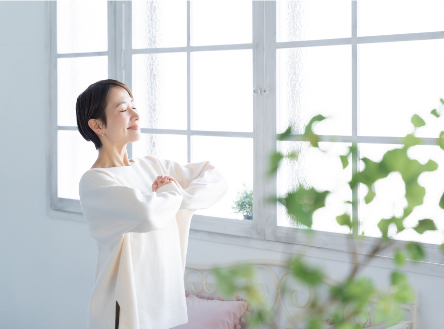 白いトップスを着た女性が自然光の差し込む窓辺で穏やかな表情を浮かべている