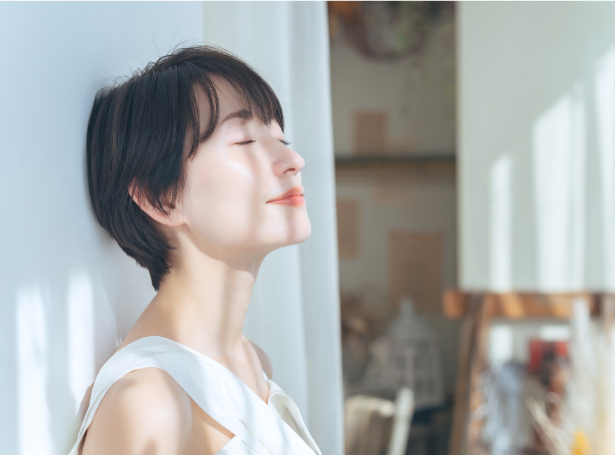 白いトップスを着た女性が自然光の差し込む窓辺で穏やかな表情を浮かべている